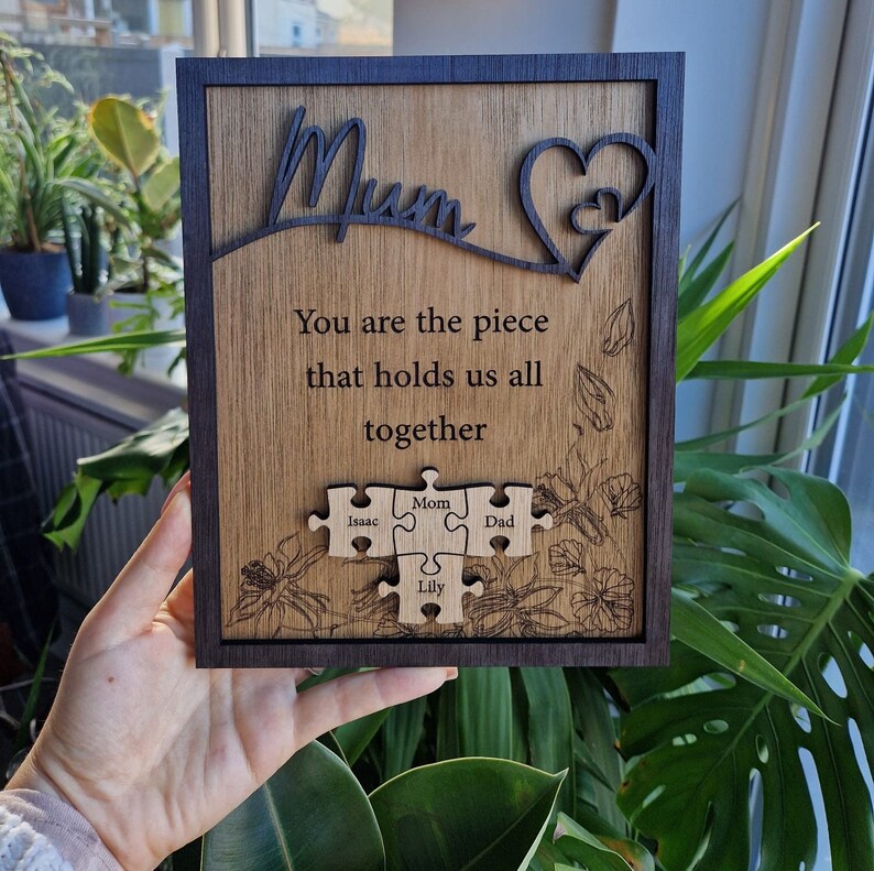 May include: Wooden plaque with a dark frame and a light wood grain center. The word "Mum" is cut out at the top, with a heart design. The text reads "You are the piece that holds us all together" with puzzle pieces below.