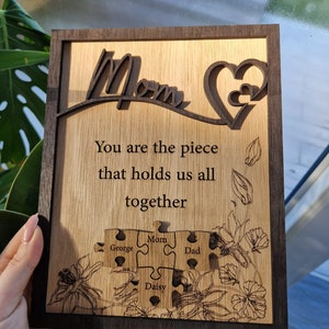 May include: Wooden plaque with a dark brown frame and a light brown interior. The plaque features the word "Mom" in a cursive font, a heart design, and the text "You are the piece that holds us all together." Below are puzzle pieces with names.