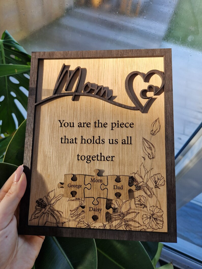 May include: Wooden plaque with a dark brown frame and a light brown interior. The plaque features the word "Mom" in a cursive font, a heart design, and the text "You are the piece that holds us all together." Below are puzzle pieces with names.