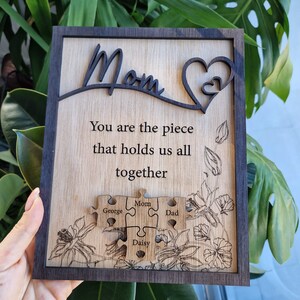 May include: Wooden plaque with a dark frame and a light wood center. The word "Mom" is cut out at the top, with a heart design. The text reads "You are the piece that holds us all together." Puzzle pieces are engraved with names.