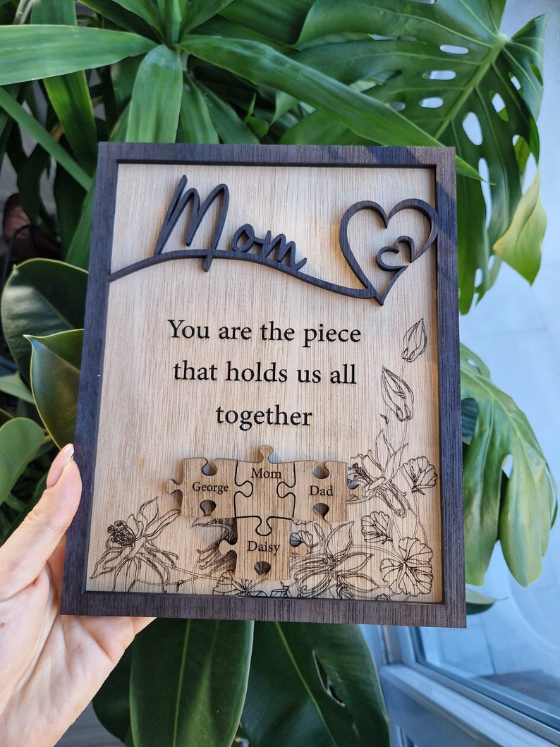 May include: Wooden plaque with a dark frame and a light wood center. The word "Mom" is cut out at the top, with a heart design. The text reads "You are the piece that holds us all together." Puzzle pieces are engraved with names.