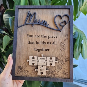 May include: Wooden plaque with the word "Mum" and a heart design. The plaque includes the text "You are the piece that holds us all together" and four puzzle pieces with the names Isaac, Mom, Dad, and Lily. The plaque has a dark brown frame.