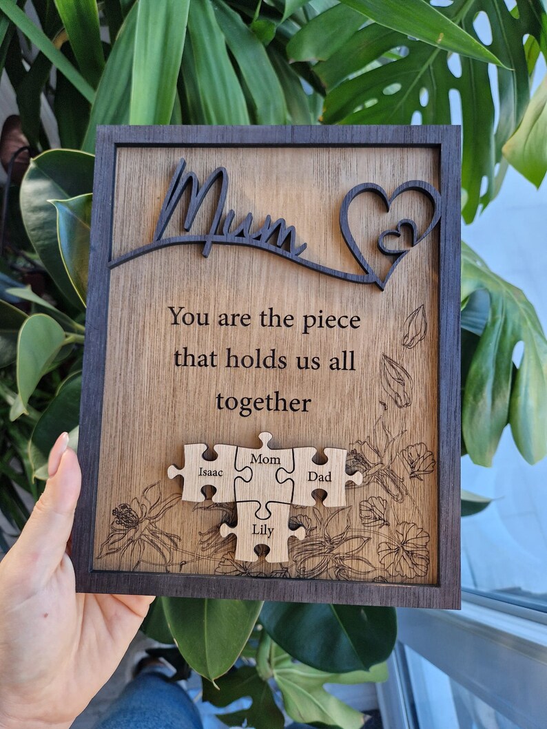 May include: Wooden plaque with the word "Mum" and a heart design. The plaque includes the text "You are the piece that holds us all together" and four puzzle pieces with the names Isaac, Mom, Dad, and Lily. The plaque has a dark brown frame.