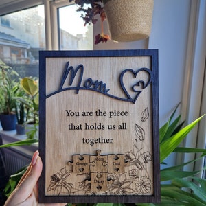 May include: Wooden plaque with a dark frame and engraved text. The plaque reads "Mom" and "You are the piece that holds us all together." It features puzzle pieces with names and floral designs. A heart shape is also included.