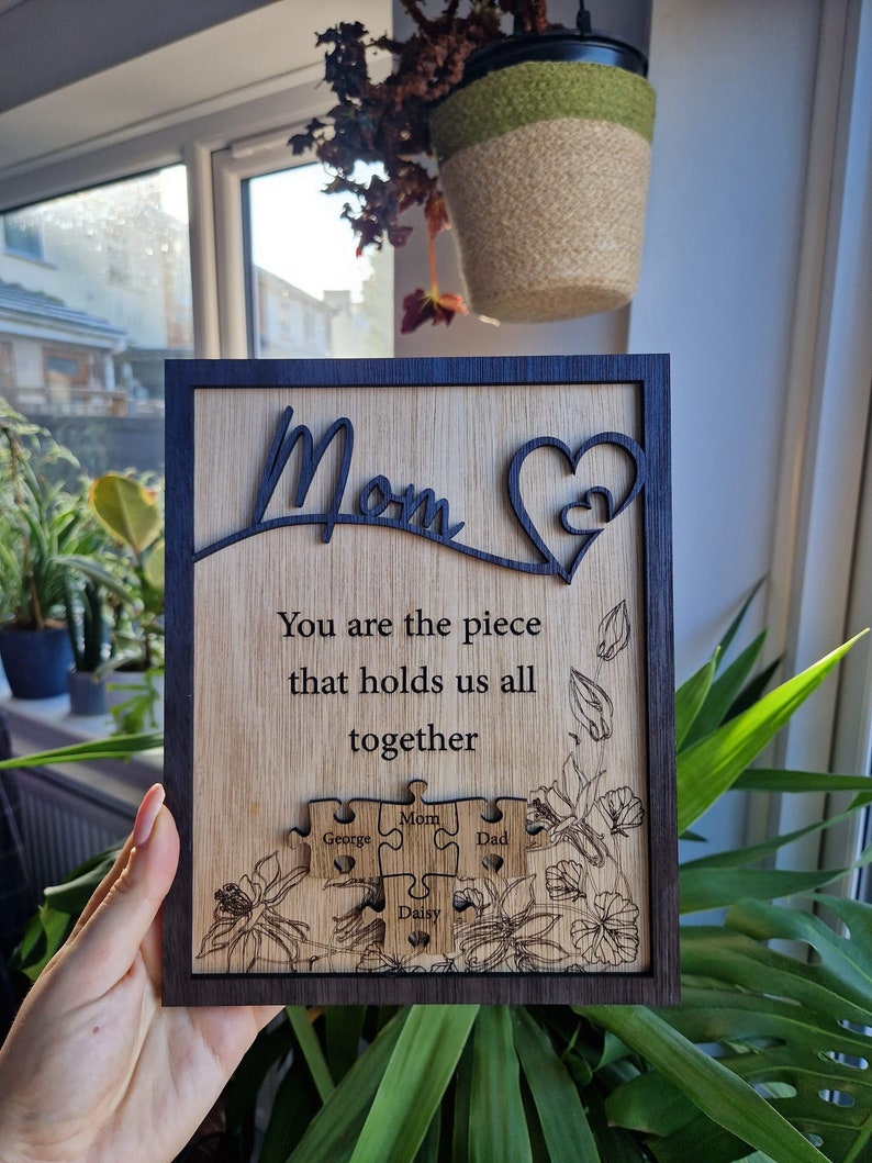 May include: Wooden plaque with a dark frame and engraved text. The plaque reads "Mom" and "You are the piece that holds us all together." It features puzzle pieces with names and floral designs. A heart shape is also included.