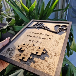May include: Wooden plaque with a dark brown frame and a light wood interior. The plaque features the word "Mom" in a cursive font, a heart design, and the quote "You are the piece that holds us all together." Four puzzle pieces are engraved with names.