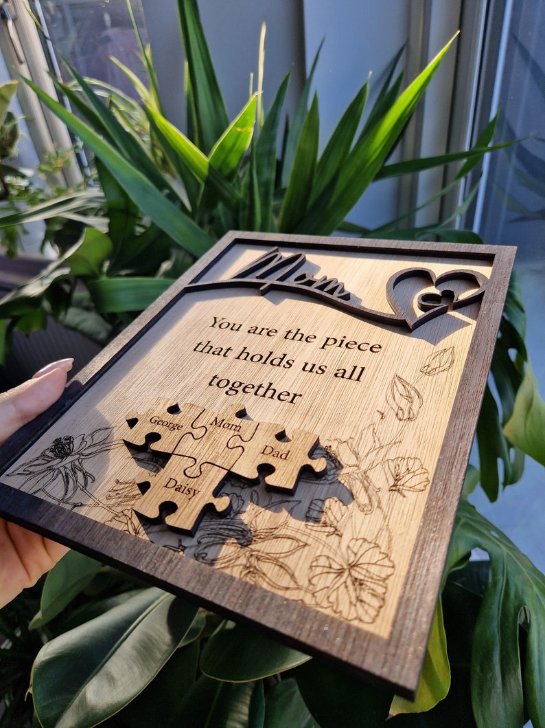 May include: Wooden plaque with a dark brown frame and a light wood interior. The plaque features the word "Mom" in a cursive font, a heart design, and the quote "You are the piece that holds us all together." Four puzzle pieces are engraved with names.