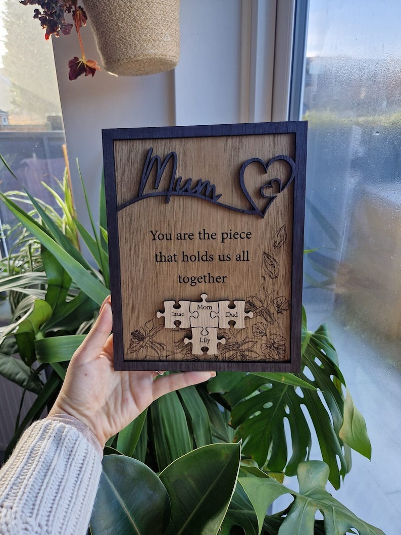 May include: Wooden plaque with a dark frame and a light wood grain center. The word "Mum" is cut out at the top, with a heart shape. The text "You are the piece that holds us all together" is engraved below. Puzzle pieces with names are at the bottom.