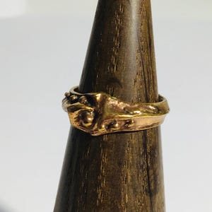 Op de afbeelding: Een goudkleurige ring met een sierlijk ontwerp, met verhoogde details en een getextureerd oppervlak. De ring wordt getoond op een houten ringhouder, die het ingewikkelde vakmanschap en de vintage esthetiek laat zien.