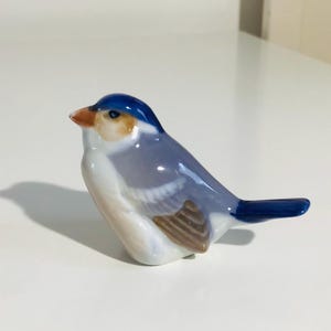 Può includere: Statuetta di uccello in ceramica con testa e coda blu, corpo azzurro e ala marrone. L'uccello ha il petto bianco e un piccolo becco arancione. La statuetta è su una superficie bianca.