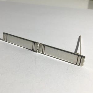 May include: A silver tie bar with a rectangular design. The tie bar has a simple, elegant style with a rectangular shape and a small, angled clip on the back. The bar is approximately 5 cm long.