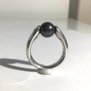 May include: A silver ring with a dark gray, spherical stone held between two curved bands. The ring is displayed on a white surface, showcasing its polished metal and the smooth texture of the stone.