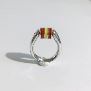 May include: A silver ring with a colorful rectangular centerpiece. The centerpiece features horizontal stripes of red, yellow, and red. The ring has a unique, curved design, and is set against a white background.