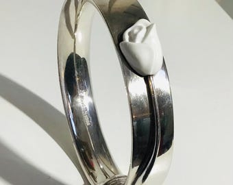 May include: A silver bangle bracelet featuring a white ceramic rose. The bracelet has a polished finish and a hinged clasp. The rose is detailed with delicate petals and a stem. A stylish piece of jewellery.