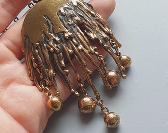 May include: A vintage brass pendant necklace. The pendant has a unique, textured design with a half-circle top and dangling, chain-connected spheres. The necklace chain is a dark bronze color, and the pendant is held in a hand.