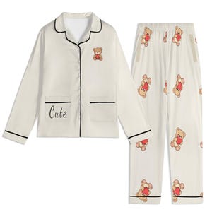 Puede incluir: Conjunto de pijama color crema con una parte superior de manga larga y pantalones. La parte superior tiene ribetes negros, un pequeño gráfico de osito de peluche y la palabra "Cute". Los pantalones presentan un patrón repetido de ositos de peluche con corazones.