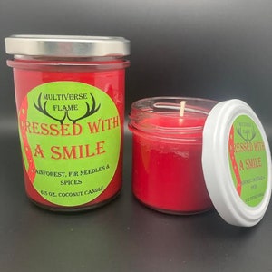 May include: Two red coconut wax candles in glass jars with silver lids. The larger candle has a green label that reads "MULTIVERSE FLAME" and "DRESSED WITH A SMILE" with the text "RAINFOREST, FIR NEEDLES & SPICES" and "6.5 OZ. COCONUT CANDLE". The smaller candle has a green label that reads "MULTIVERSE FLAME" and "DRESSED WITH A SMILE" with the text "RAINFOREST, FIR NEEDLES & SPICES" and "3.12 OZ. COCONUT CANDLE".