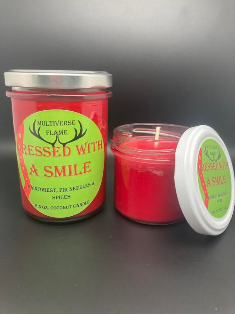 May include: Two red coconut wax candles in glass jars with silver lids. The larger candle has a green label that reads "MULTIVERSE FLAME" and "DRESSED WITH A SMILE" with the text "RAINFOREST, FIR NEEDLES & SPICES" and "6.5 OZ. COCONUT CANDLE". The smaller candle has a green label that reads "MULTIVERSE FLAME" and "DRESSED WITH A SMILE" with the text "RAINFOREST, FIR NEEDLES & SPICES" and "3.12 OZ. COCONUT CANDLE".