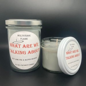 May include: Two glass jars with white lids and labels. The labels are white with black text and a red circle that says "What are we talking about". The text on the labels says "Multiverse Flame" and "Key Lime Pie & Blood Orange 6.5 oz. Coconut Candle".