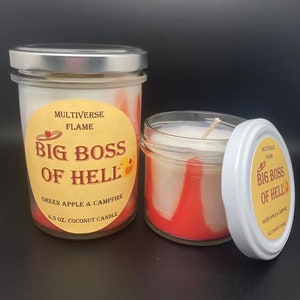 May include: Two glass jars with red and white wax candles. The jars have labels that say "MULTIVERSE FLAME BIG BOSS OF HELL GREEN APPLE & CAMPFIRE 6.5 OZ. COCONUT CANDLE".
