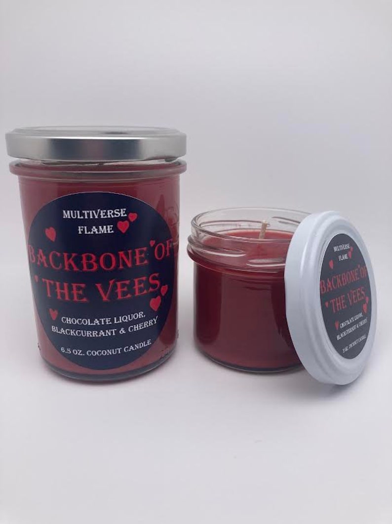 May include: Two red glass jars with silver lids. The jars contain red wax and a wick. The jars have a label that reads "MULTIVERSE FLAME BACKBONE OF THE VEES CHOCOLATE LIQUOR BLACKCURRANT & CHERRY 6.5 OZ. COCONUT CANDLE".