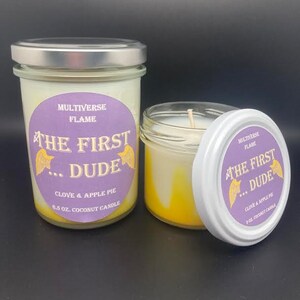 May include: Two glass jars with lids containing coconut candles. The jars have purple labels with the text "MULTIVERSE FLAME THE FIRST...DUDE CLOVE & APPLE PIE 6.5 OZ. COCONUT CANDLE" and "MULTIVERSE FLAME THE FIRST...DUDE CLOVE & APPLE PIE 3 OZ. COCONUT CANDLE".
