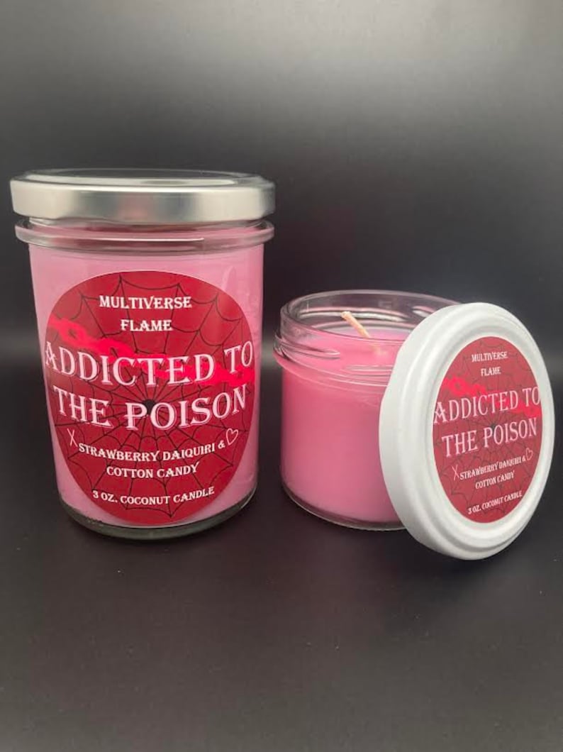 May include: Two pink glass jars with silver lids. The jars contain a pink candle and have a red and white label that reads "Addicted to the Poison" with the scent "Strawberry Daiquiri & Cotton Candy". The label also says "3 oz. Coconut Candle" and "Multiverse Flame".