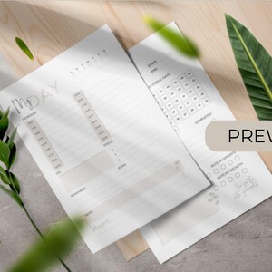 Könnte beinhalten: Zwei weiße Seiten eines Tagesplaners mit minimalistischem Design. Eine Seite zeigt ein Zeitplan-Layout mit Zeitfenstern, die andere enthält einen Gewohnheitstracker. Das Wort "PREVIEW" ist auf der rechten Seite sichtbar. Grüne Blätter und eine Holzoberfläche im Hintergrund.