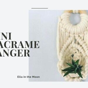 以下が含まれることがあります： 木製の輪っかが付いた白いマクラメハンガー。ハンガーは結び目の技術で作られており、小さな植物が入っています。白い背景に黒い文字で「MINI MACRAME HANGER」と書かれています。白い背景に黒い文字で「Ella in the Moon」と書かれています。