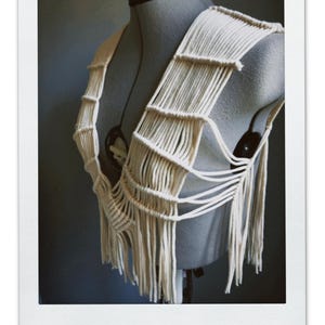 以下が含まれることがあります： マネキンに展示されたクリーム色のマクラメトップ。手作りの衣服は、垂直のフリンジが付いた織りデザインが特徴です。このトップは、複雑な結び目技術とボヘミアンな美学を披露する、ユニークなウェアラブルアートです。