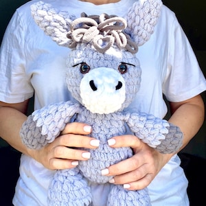 May include: A gray crocheted donkey plush toy with brown yarn for the mane and tail. The donkey has large brown eyes and a white muzzle.