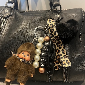 Puede incluir: Bolso de mano de cuero negro con herrajes plateados y cierre de cremallera. Un llavero con un mono de peluche marrón, cuentas de perlas y el nombre "BELLA" deletreado en cuentas de letras negras. Una cinta con estampado de leopardo y un corazón negro y esponjoso completan los accesorios.