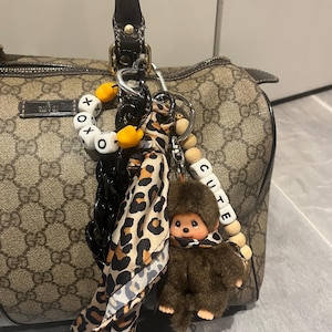 Puede incluir: Un bolso de mano marrón y beige con un patrón de logotipo repetido. Se adjuntan varios accesorios, incluyendo un pequeño mono de peluche, una bufanda con estampado de leopardo y abalorios con las palabras "cute" y "xoxo".