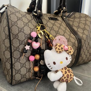 Może przedstawiać: Brązowa i czarna torebka z wzorem, z złotym łańcuszkiem i brelokiem Hello Kitty. Brelok przedstawia białego Hello Kitty w brązowo-czarnym stroju w panterkę.