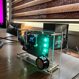 May include: A clear acrylic case containing a black circuit board. The board features a digital display showing "810" and a green LED light. A black knob is located on the bottom right corner of the board.