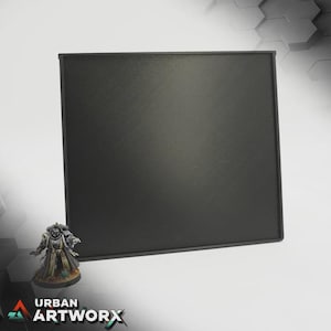 Puede incluir: Una alfombra de juego cuadrada y negra con una superficie texturizada. Una figura en miniatura está colocada a la izquierda de la alfombra. El logotipo de Urban Artworx está en la esquina inferior izquierda.