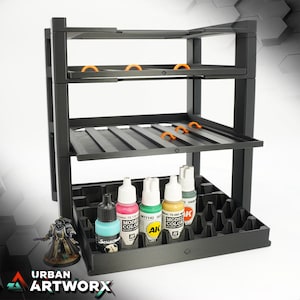 Può includere: Un rack per vernici in plastica nera con tre ripiani e un vassoio rimovibile. Il rack è progettato per contenere piccoli flaconi di vernice. Il vassoio ha una griglia di fori per mantenere i flaconi in posizione verticale. Ci sono cinque flaconi di vernice nel vassoio, e sono di vari colori, tra cui verde, rosso e oro. Il rack è etichettato "Urban Artworx".