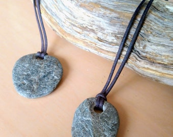 May include: Two gray stone pendants on brown leather cords. The stones are smooth and have a natural, irregular shape. The cords are tied together at the top.