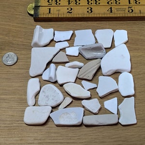 Puede incluir: Una colección de trozos de vidrio de mar en varias formas y tonos de blanco, beige y gris, dispuestos sobre una superficie de madera. Una regla de madera vintage, que mide hasta 89 cm, y una moneda también están presentes.