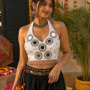 Puede incluir: Un top blanco tipo halter con adornos de espejos circulares y un dobladillo con flecos de cuentas, combinado con una falda negra que tiene una cintura decorativa con bordados coloridos, espejos pequeños y borlas de tela colgantes.