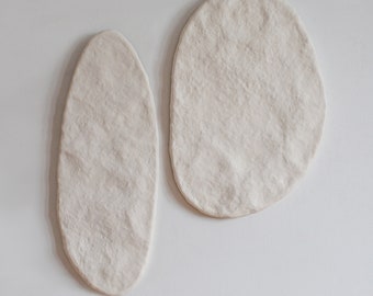 Könnte beinhalten: Zwei cremefarbene, strukturierte Wandkunstwerke in organischer Form. Eines ist länglich und oval, das andere hat eine größere, abgerundete Form. Die Stücke sind an einer weißen Wand befestigt und schaffen eine minimalistische, moderne Ästhetik.