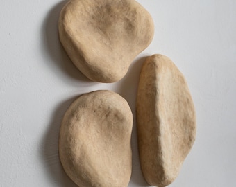 Könnte beinhalten: Drei abstrakte Wandskulpturen in einem neutralen Beige. Die Formen sind organisch und unregelmäßig und ähneln glatten Steinen oder Kieselsteinen. Die Skulpturen sind an einer weißen Wand angebracht und schaffen eine minimalistische und moderne Ästhetik.