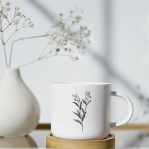 May include: White ceramic mug with a floral design featuring a sprig of lavender-colored flowers and green leaves. The mug sits on a round, light-colored wooden coaster. A white vase with delicate white flowers is in the background.