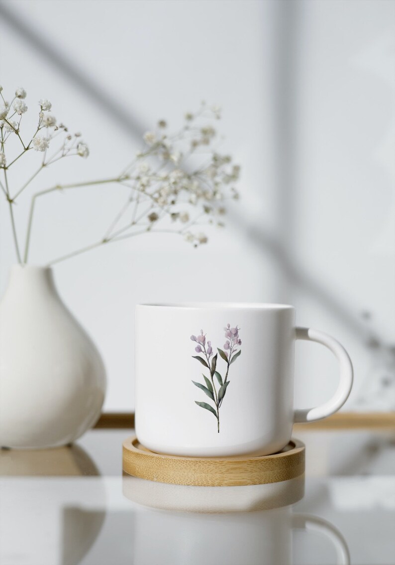 May include: White ceramic mug with a floral design featuring a sprig of lavender-colored flowers and green leaves. The mug sits on a round, light-colored wooden coaster. A white vase with delicate white flowers is in the background.