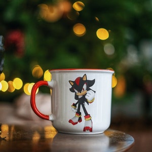May include: White ceramic coffee mug with a red rim. The mug features a black and red cartoon character with a red and yellow design. The mug is sitting on a wooden table.