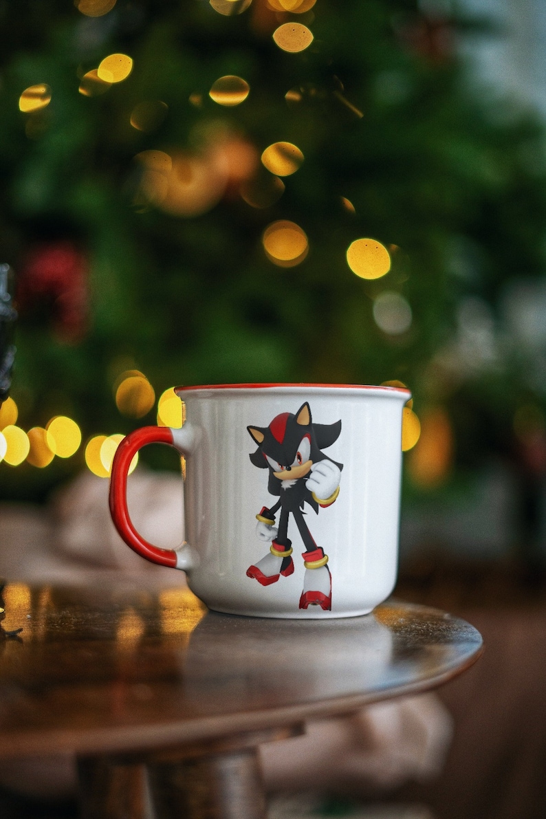 May include: White ceramic coffee mug with a red rim. The mug features a black and red cartoon character with a red and yellow design. The mug is sitting on a wooden table.