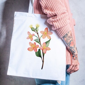 May include: White tote bag featuring a watercolor illustration of a flowering branch with peach-colored blossoms, green leaves, and yellow accents. The bag is held against a background of a pink sweater and blue jeans.