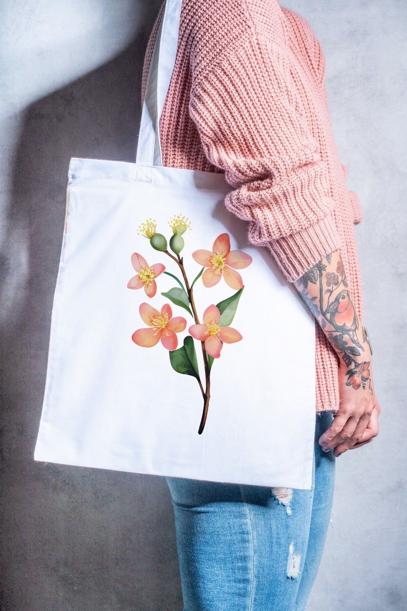 May include: White tote bag featuring a watercolor illustration of a flowering branch with peach-colored blossoms, green leaves, and yellow accents. The bag is held against a background of a pink sweater and blue jeans.