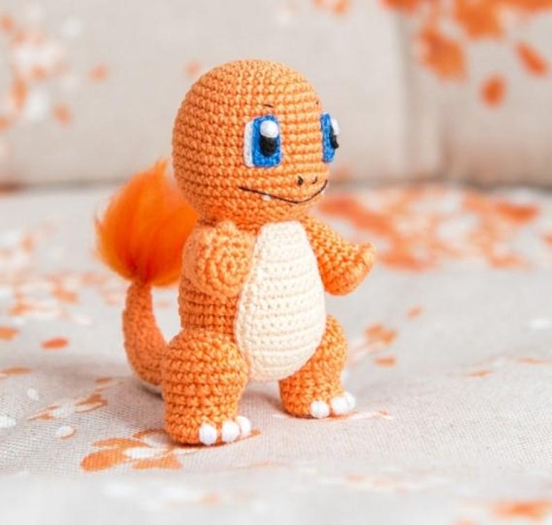 May include: A crocheted orange and white plush toy resembling a cartoon character with a tail. The toy has blue eyes and a smiling expression.