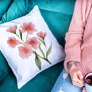 May include: White square pillow with a watercolor floral design featuring pink flowers and green leaves. The pillow is placed on a teal couch. A person wearing a pink sweater is holding a mug of a dark beverage.